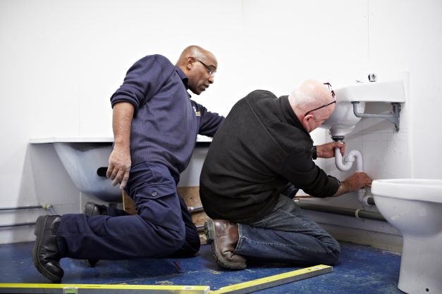 Blog post image for 'First steps to becoming a qualified plumber' shows plumbing tutor assisting a student with fitting a waste pipe to a basin in a bathroom