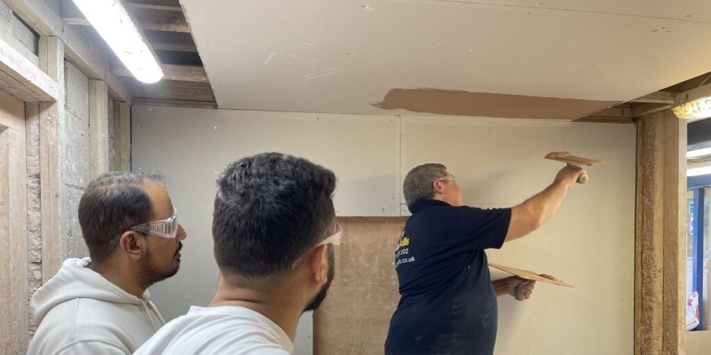 An Able Skills plastering instructor is showing students how to apply plaster to a ceiling. Two students are learning plastering. They are turned towards the instructor watching the demonstration.