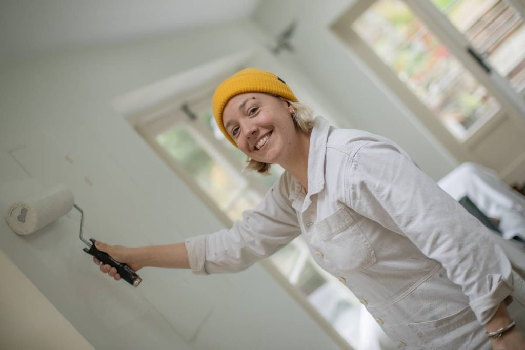 Photo of Katie Corkill using a paint roller, applying paint to a wall. She is wearing white overalls and a yellow hat.