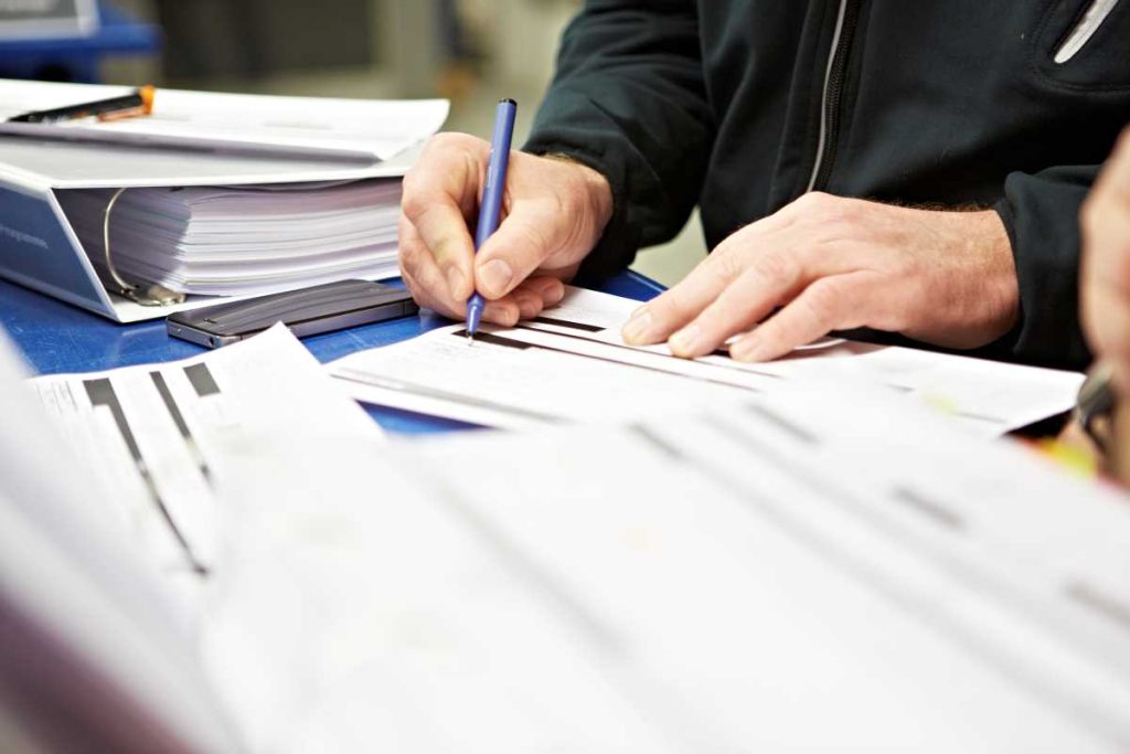 Person holding a pen and writing in a training booklet. There are other sheets of paper and a lever arch folder on the desk. 