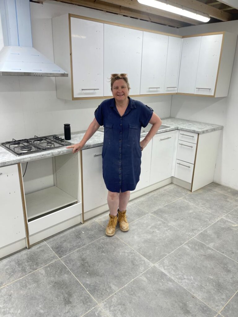 Pam is standing in front of fitted kitchen units wearing work boots and a blue dress.